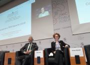 Allan Rocha and Teresa Hackett participating in a working session at the WTO Public Forum. They are seated, in front of a large screen where information / visuals are projected.