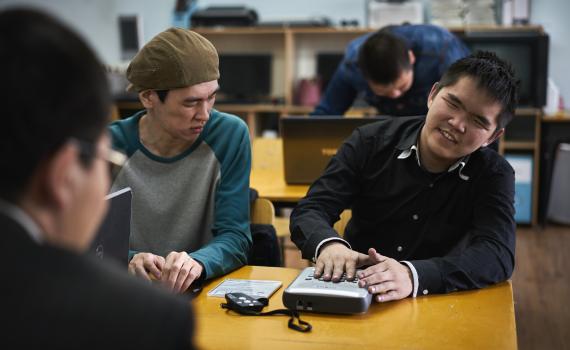 Two young men sitting at a library desk working with new Daisy players. The instruction booklet is on the desk.